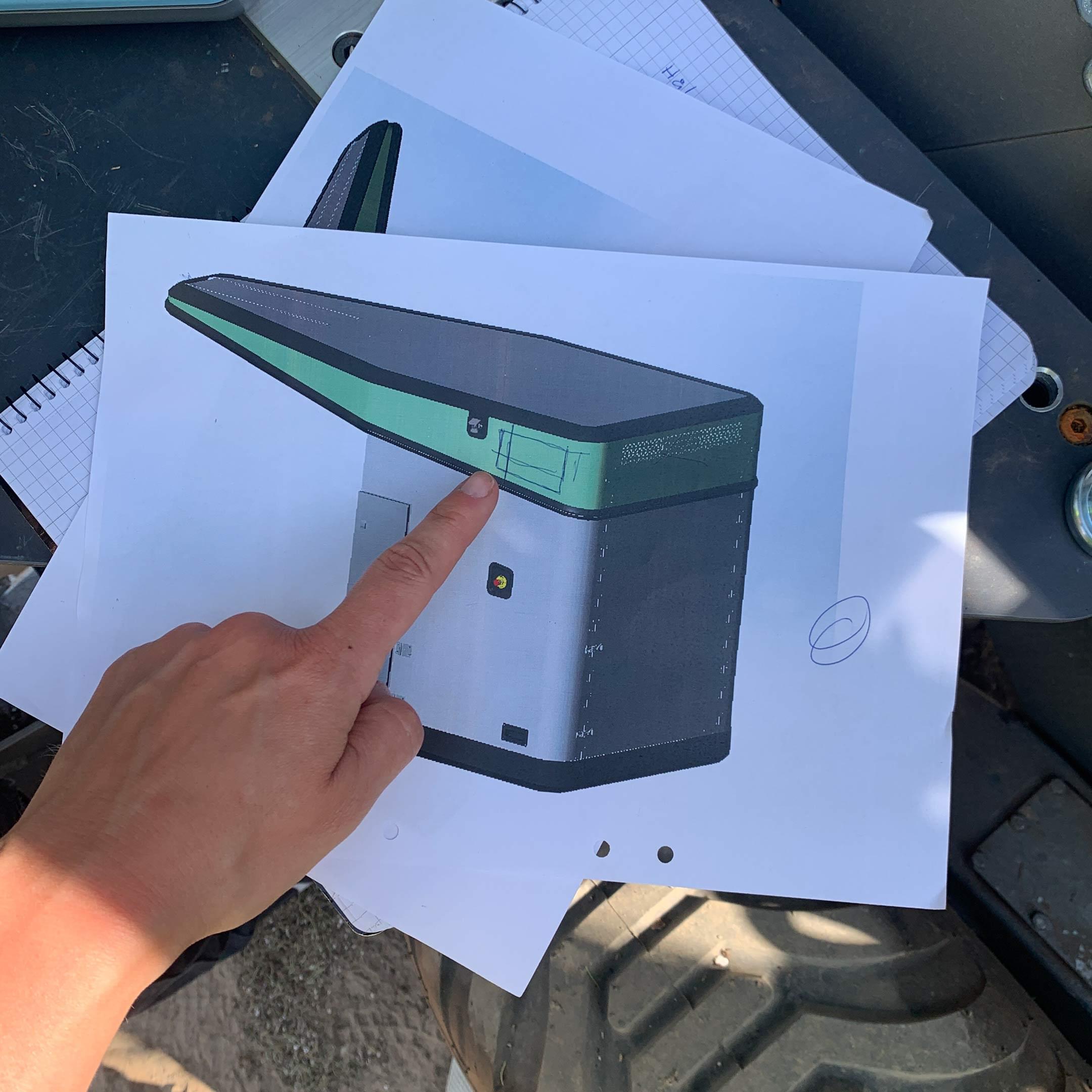 Finger som pekar på en utskriven skiss av planteringsroboten som ligger på den fysiska robotens däck