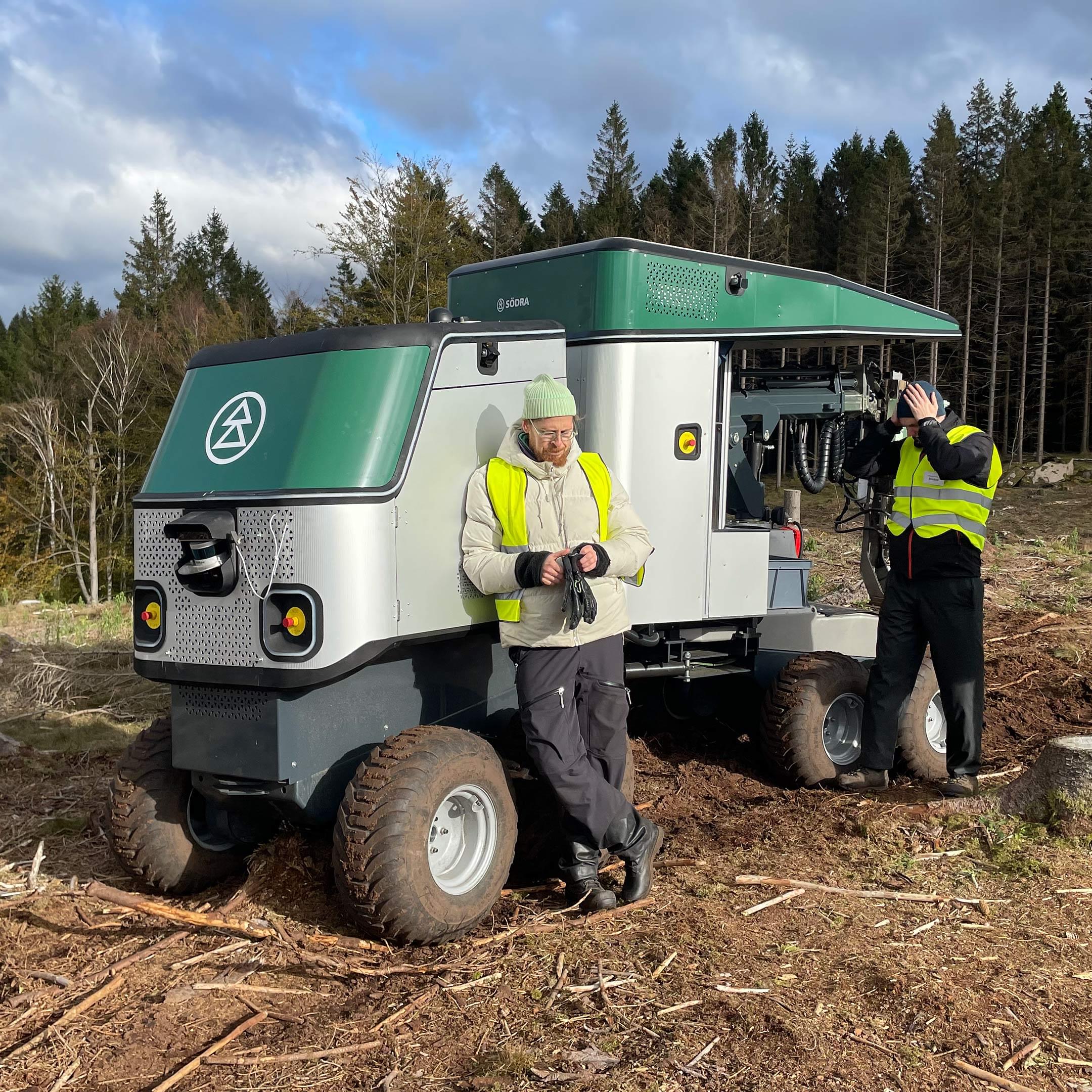 Två personer i reflexväst som står framför planteringsroboten i skogen.